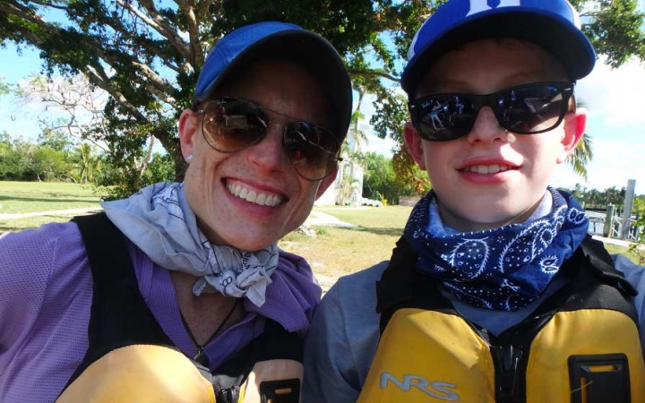 A parent and child wearing life jackets, sunglasses and hats smile at the camera.